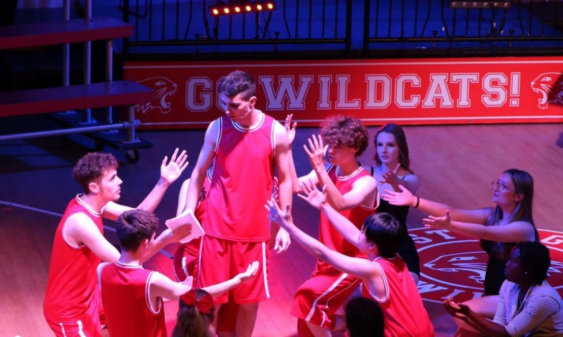 Pupils from Kennet School perform a dynamic basketball team scene during their high school musical production, dressed in team uniforms mid-action on 